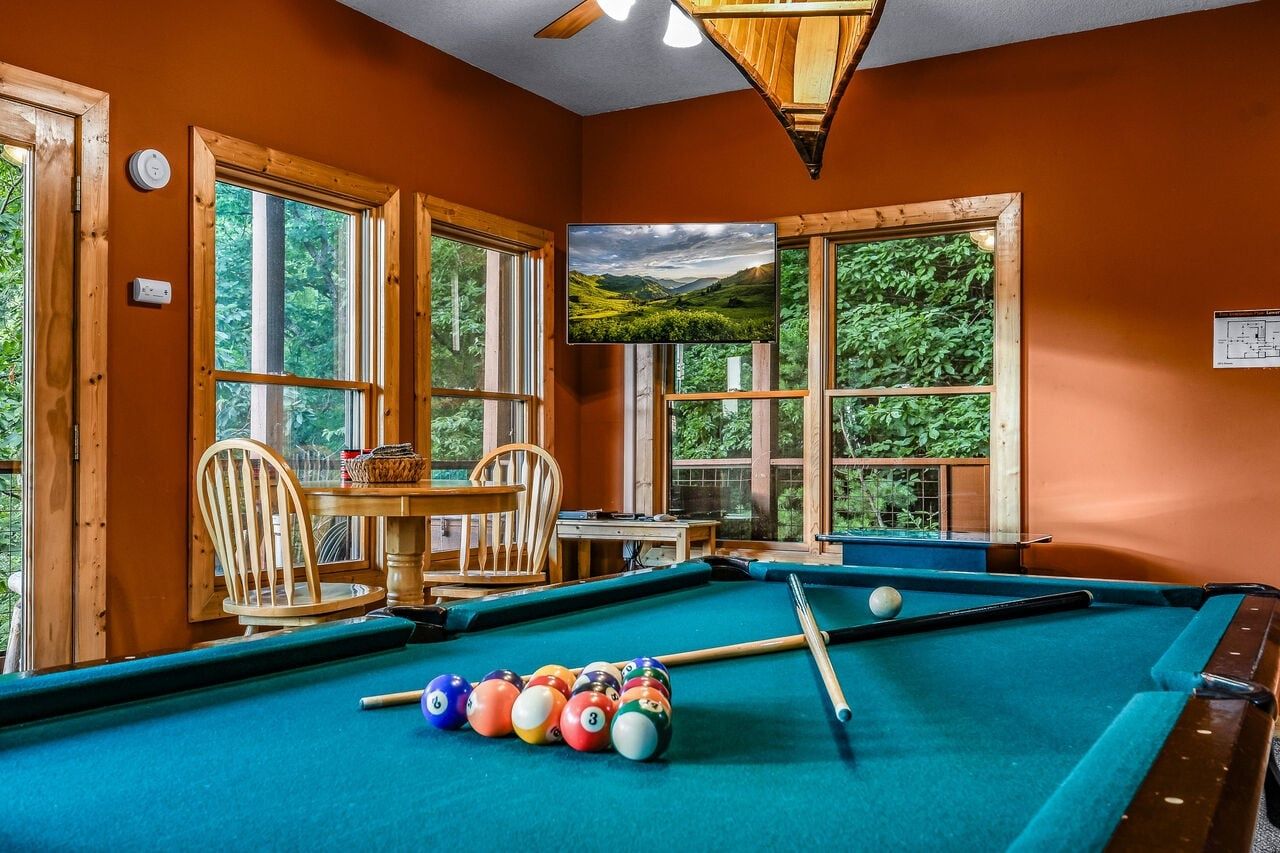 A pool table with balls, a chair, a table with a basket, and a television displaying a landscape.