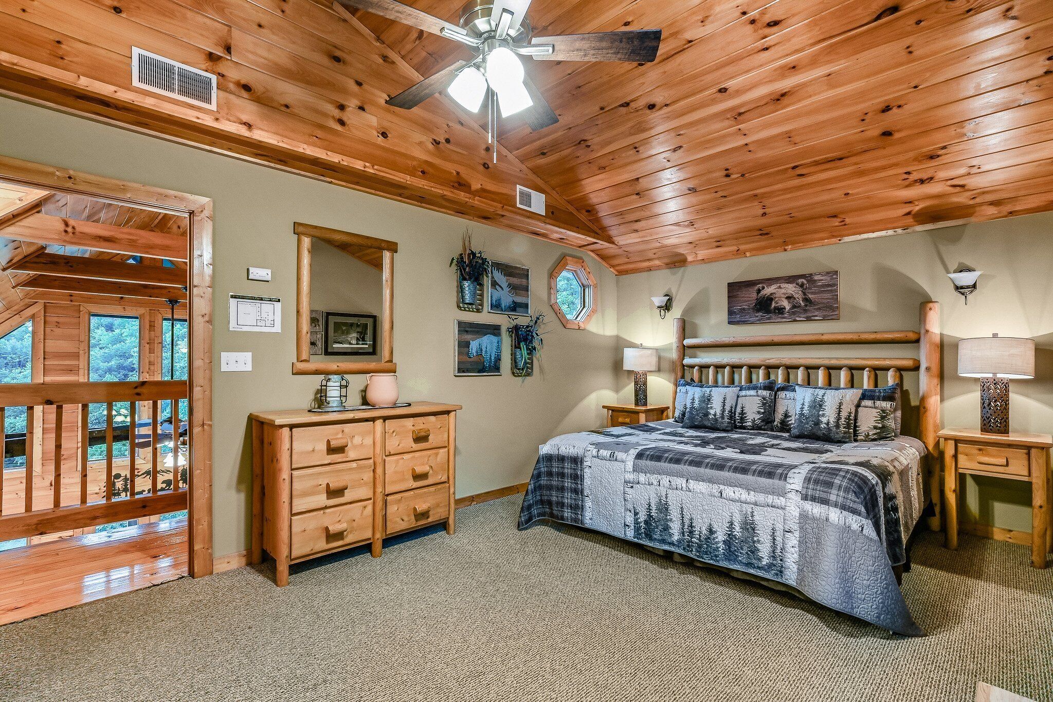 A bedroom with a wooden ceiling, a bed with a forest-themed bedspread, a wooden dresser, a nightstand with a lamp, and a window with a view of the outdoors.