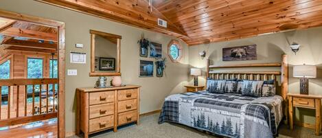 A bedroom with a wooden ceiling, a bed with a forest-themed bedspread, a wooden dresser, a nightstand with a lamp, and a window with a view of the outdoors.