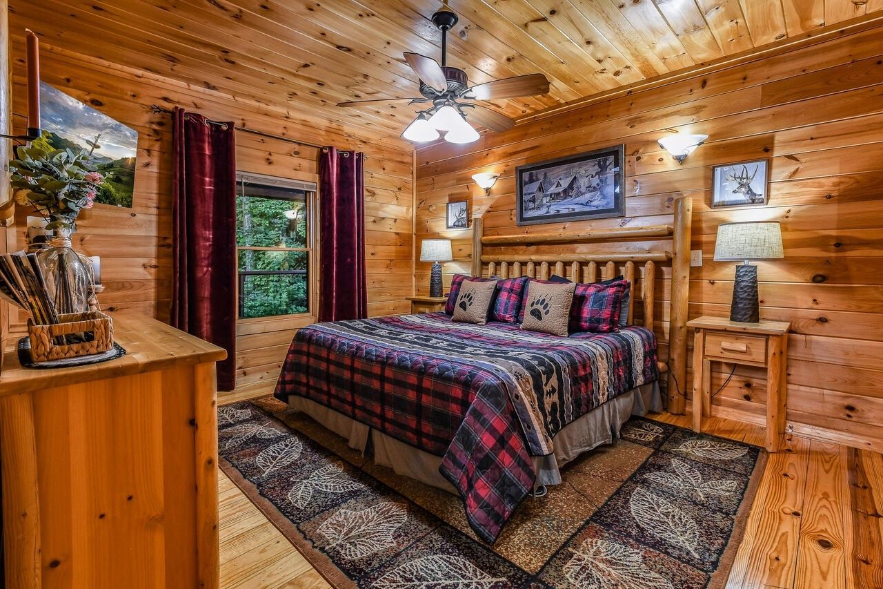 Une chambre à coucher dans une cabane en bois, avec un lit, une table de chevet, un ventilateur de plafond et une fenêtre avec des rideaux.