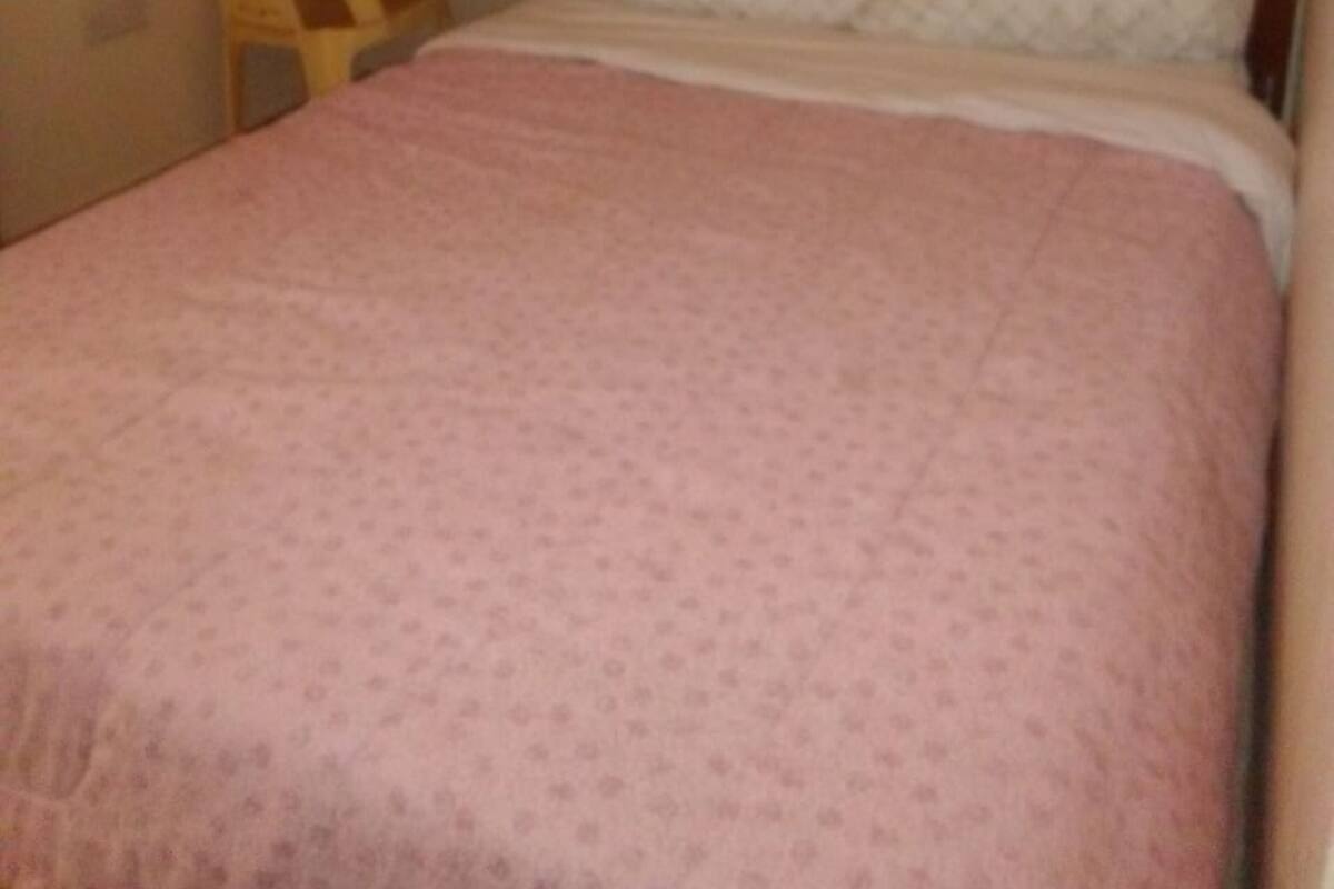 Uma cama de solteiro com colcha rosa, mesa de cabeceira de madeira e uma cadeira.