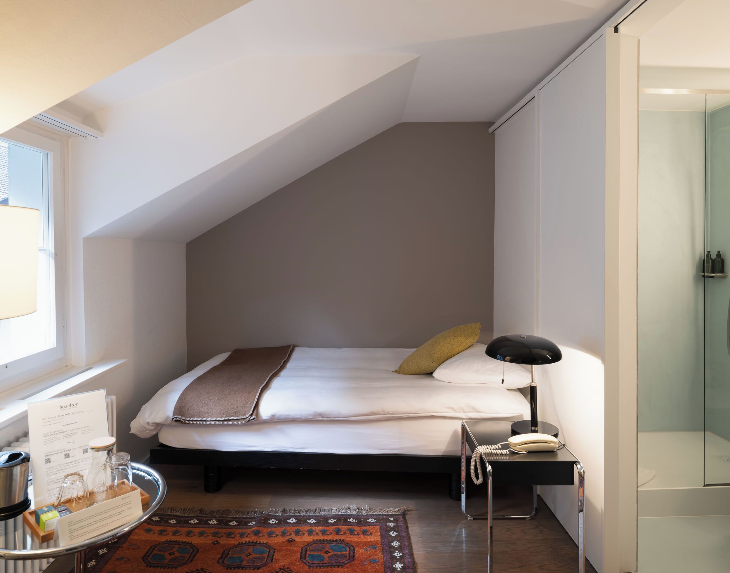 A modern bedroom with a sloped ceiling, a bed with white linens, a bedside table with a lamp, a glass shower enclosure, and a small rug.