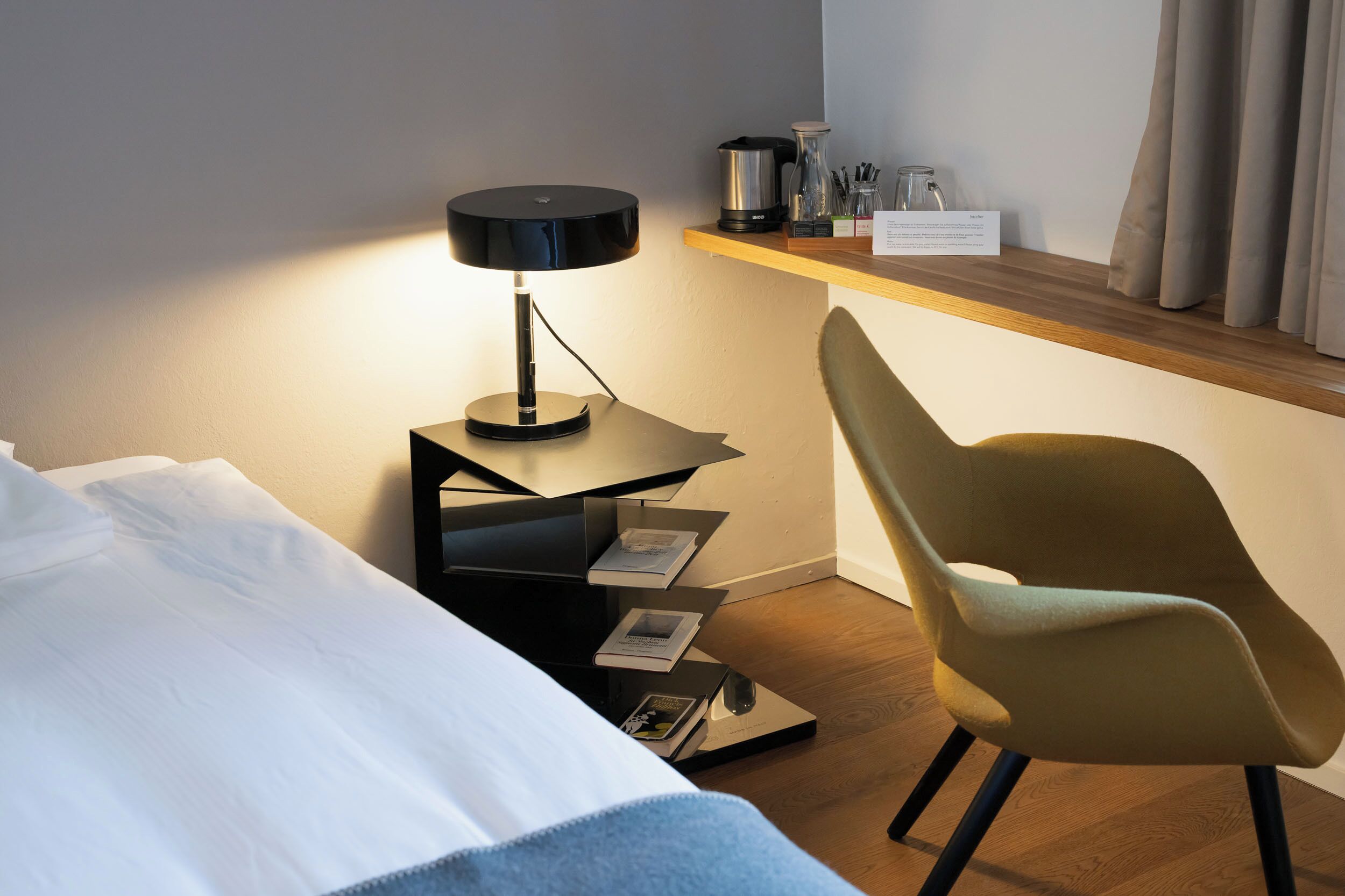 Habitación de hotel ordenada con una cama, un escritorio con lámpara, una silla y un estante con varios objetos.