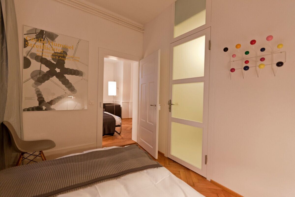 A hotel room with a bed, a chair, a wall art piece, a door with frosted glass panels, and a wall-mounted color dot display.
