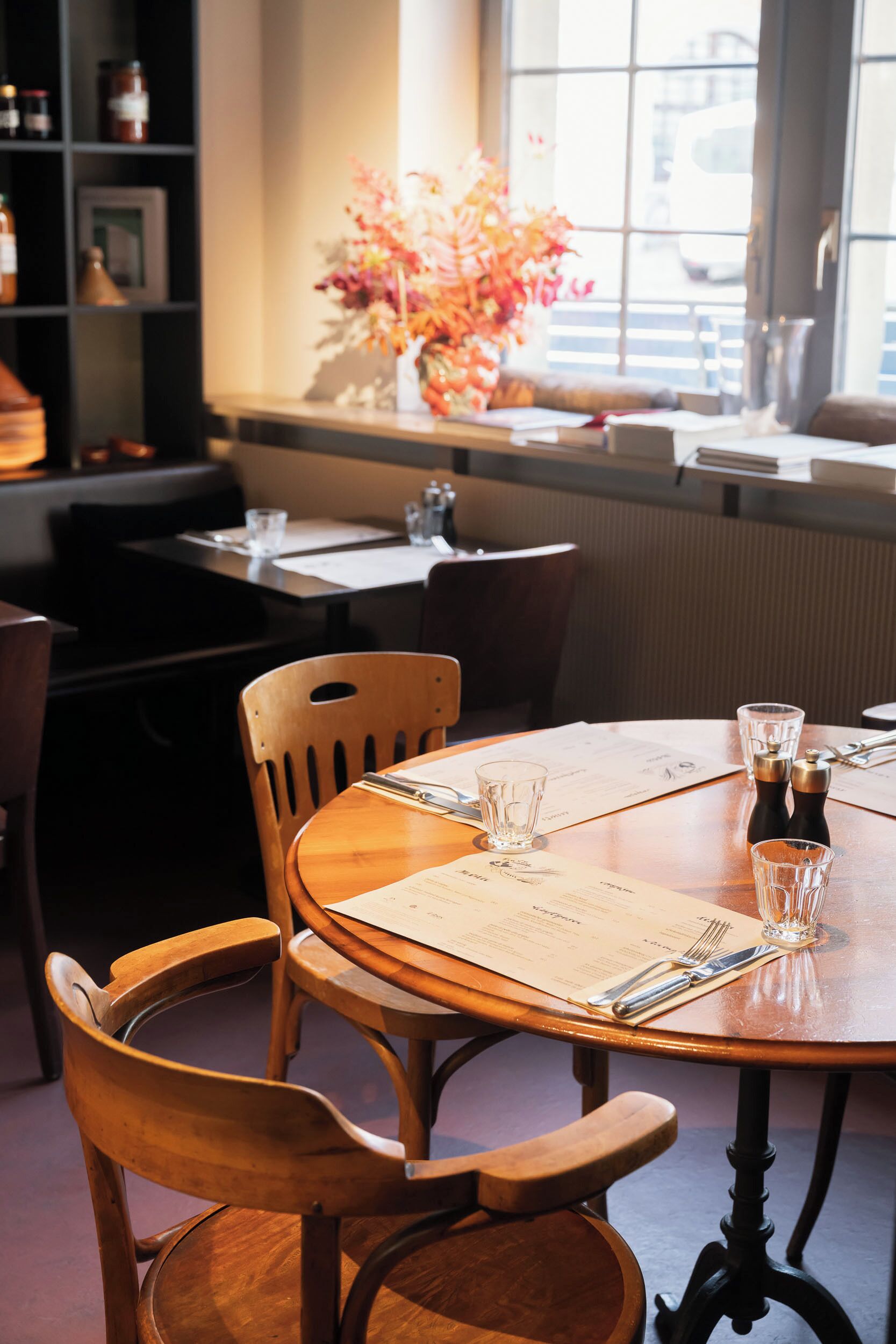 A dining area with wooden chairs and tables, a menu, salt and pepper shakers, and a vase with flowers.