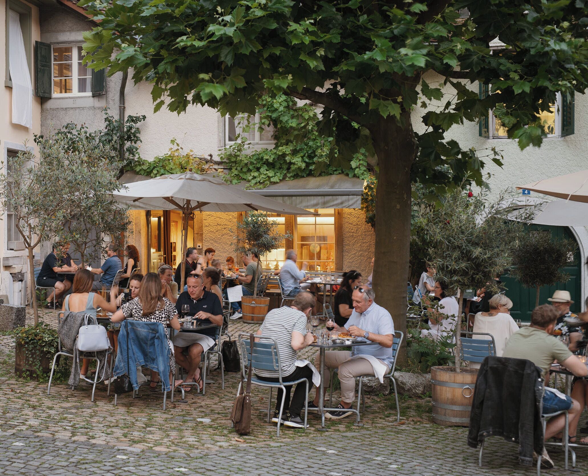 En udendørs café omgivet af træer og bygninger, med borde og stole.