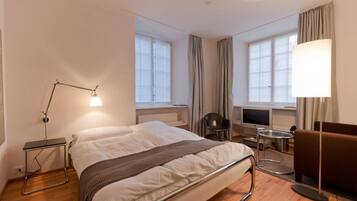 A hotel room with a bed, bedside table, lamp, chair, TV, and windows.