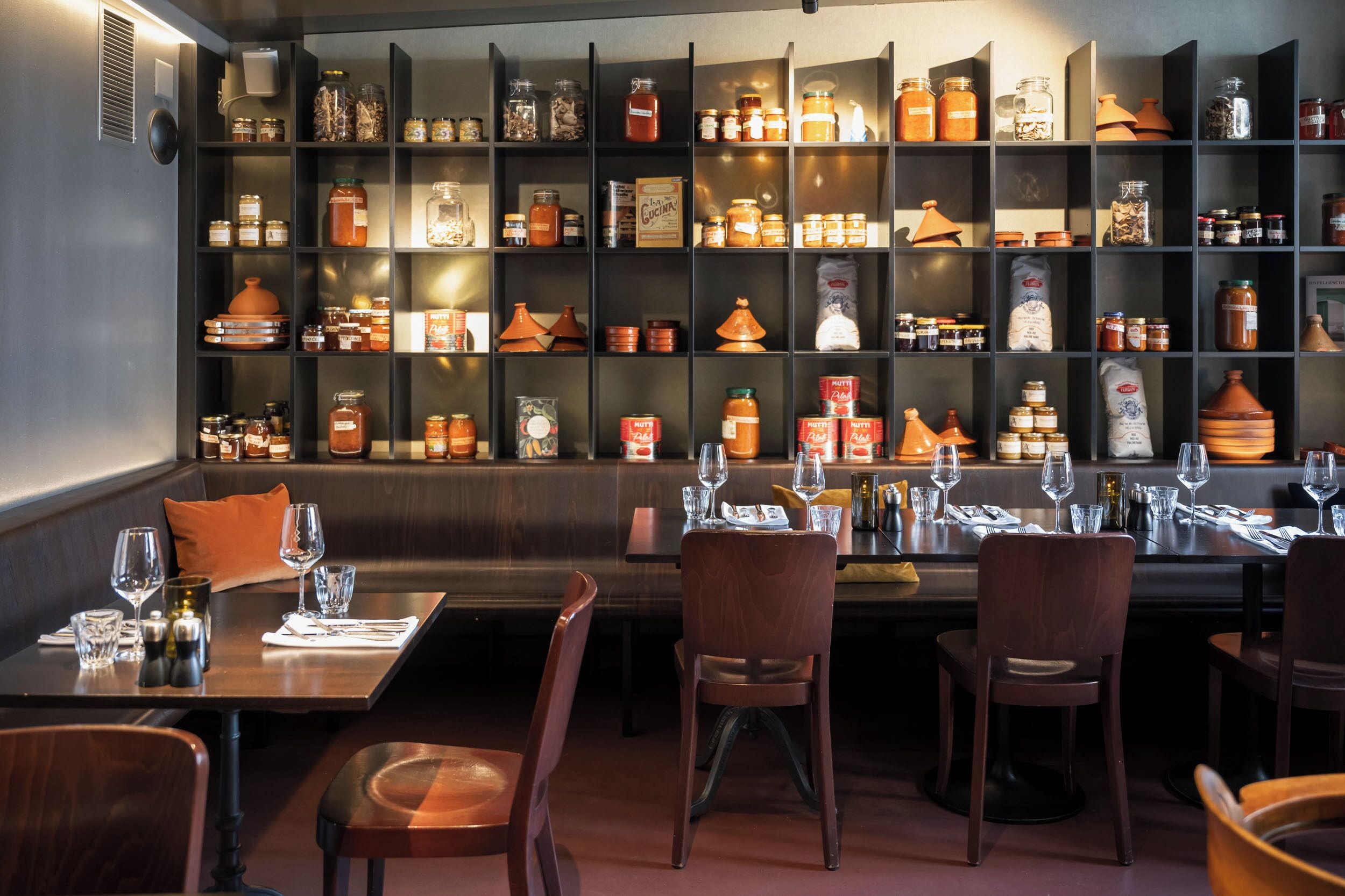 Ein Restaurantinneres mit mehreren Esstischen, Stühlen und Regalen, die mit Gläsern und Küchenutensilien gefüllt sind.