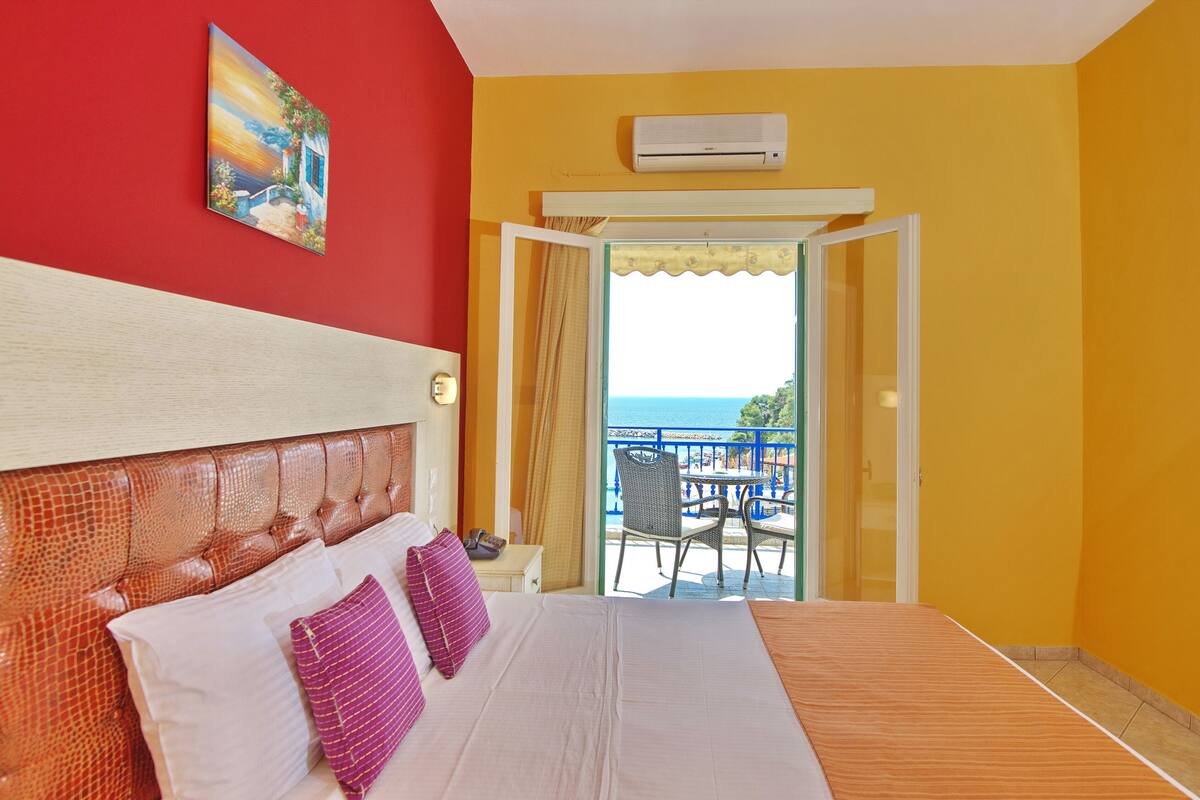 A hotel room with a large bed, a painting on the wall, an air conditioning unit, and a balcony with a view of the sea.