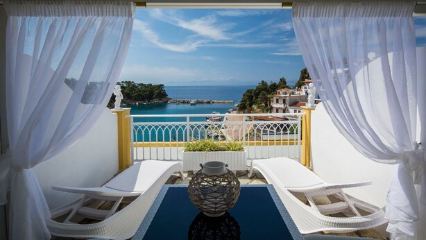 A balcony with white wicker chairs and a table, overlooking a coastal view with boats and buildings. - Pension Votsi (Alonissos)