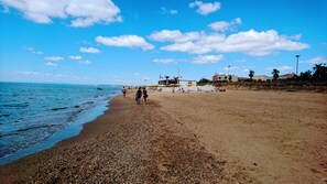 A pebbly beach with a clear sky, a few people, and a building with a flag in the background.