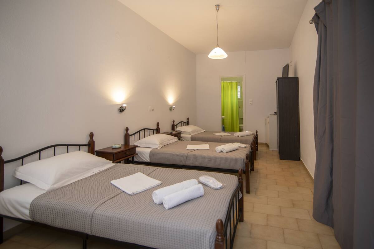 Habitación con tres camas individuales, cada una con cabecera de madera, una mesita de noche y una lámpara. Se observa un cortinaje verde en la puerta, y un mueble oscuro apoyado en la pared.