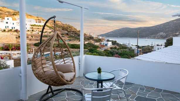 A balcony with a wicker chair, a small table, and a view of a coastal town and sea. - Prasini Gonia (Sifnos)