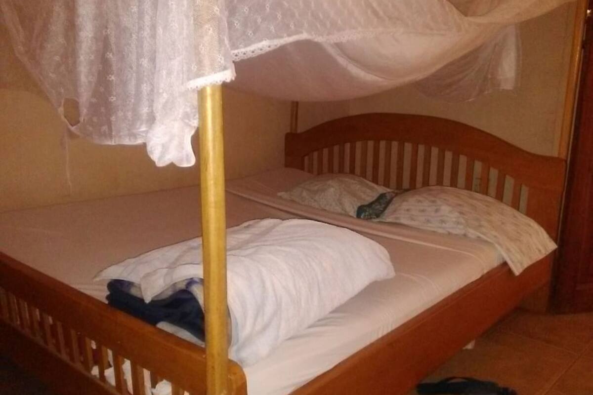 A single bed with a wooden frame and a canopy, a pair of slippers on the floor, and a plain wall.