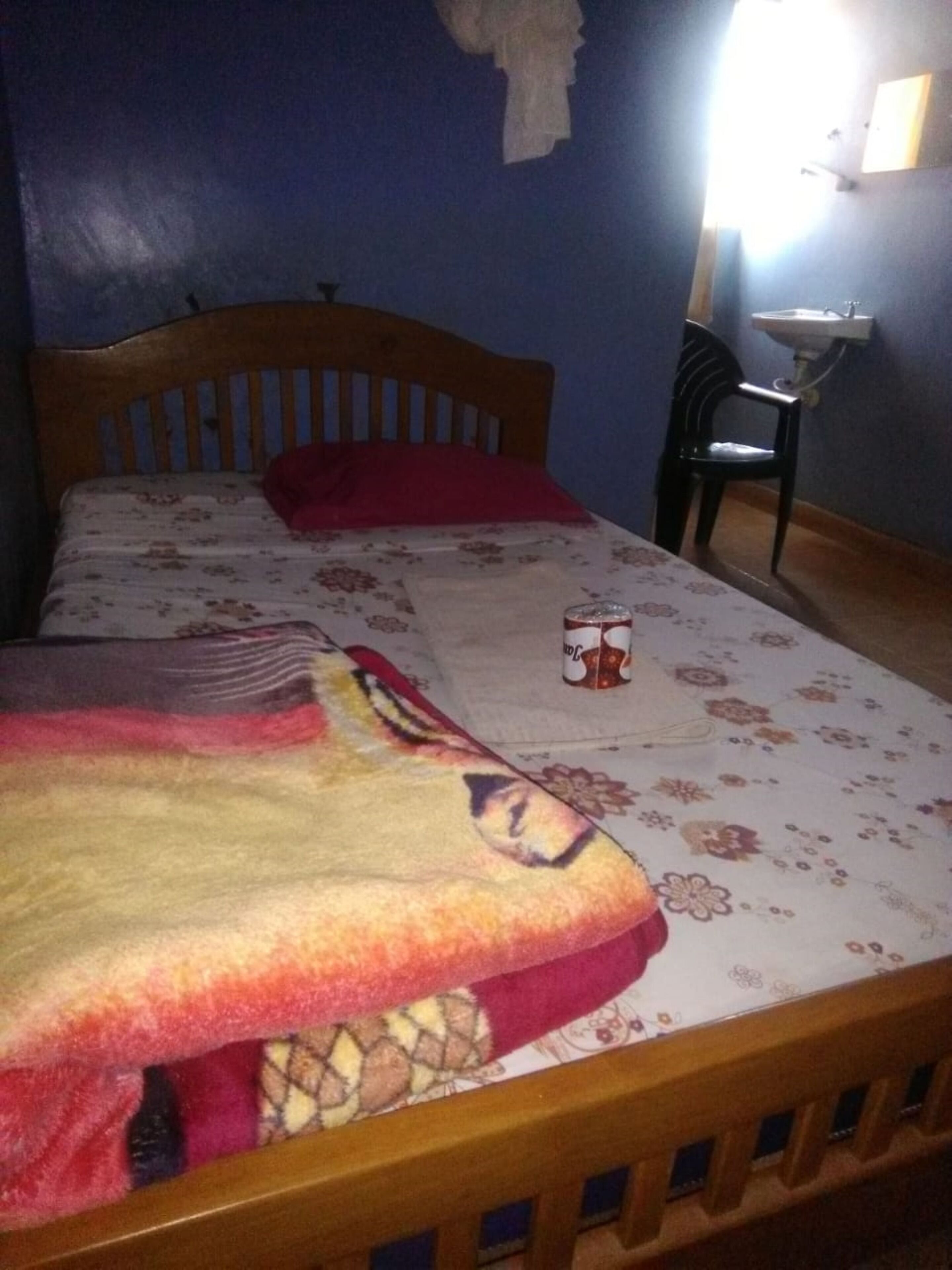A bedroom with a wooden bed, a red pillow, a floral bedspread, a small bedside table with a lamp, and a chair.