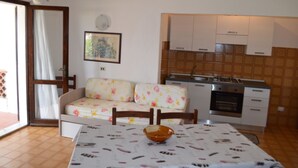A cozy living room with a sofa and a dining table, a kitchen with cabinets and appliances, and a door leading outside. - THREE-ROOM APARTMENT BADAS Costa Rei Centro (Costa Rei)
