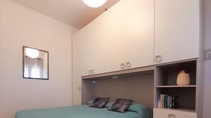 A bedroom with a bed, a nightstand, a dresser, and a shelf with books. - THREE-ROOM APARTMENT BADAS Costa Rei Centro (Costa Rei)