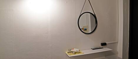 A bathroom with a white wall, a hanging mirror, a shelf with toiletries, and a light fixture.