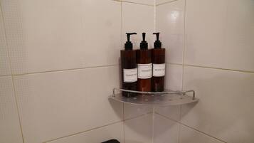 Un baño con paredes de azulejos blancos, un taburete negro y un estante con tres botellas etiquetadas como 'Hand Wash' y 'Body Wash'.