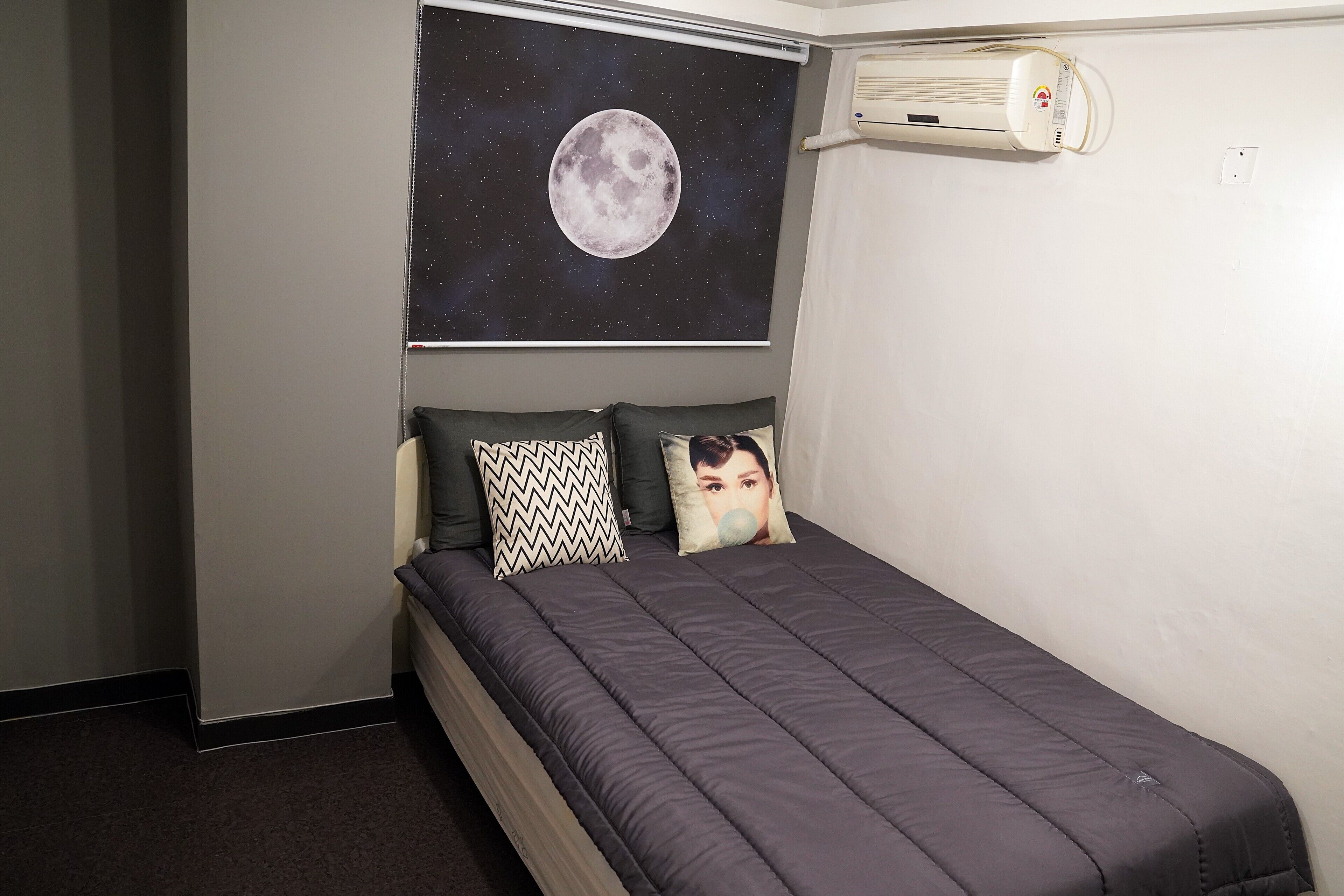 A single bed with a dark quilt, two patterned pillows, and a wall-mounted air conditioner.