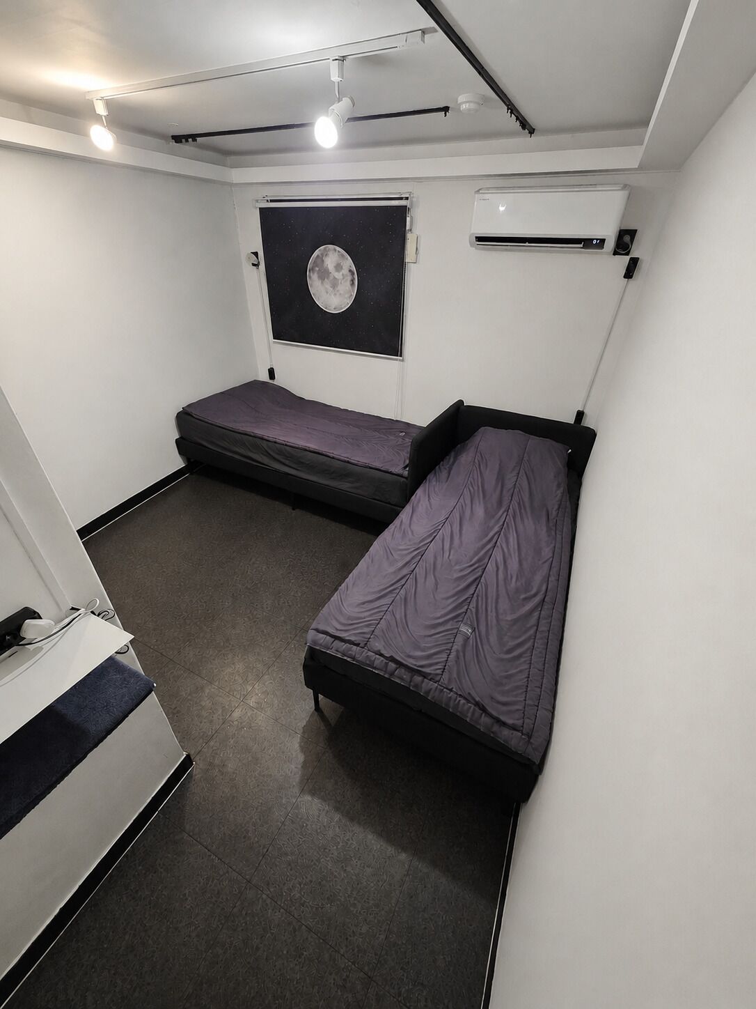 Habitación pequeña con dos camas individuales, ventilador de techo, equipo de aire acondicionado y ventana con un cartel de la luna.