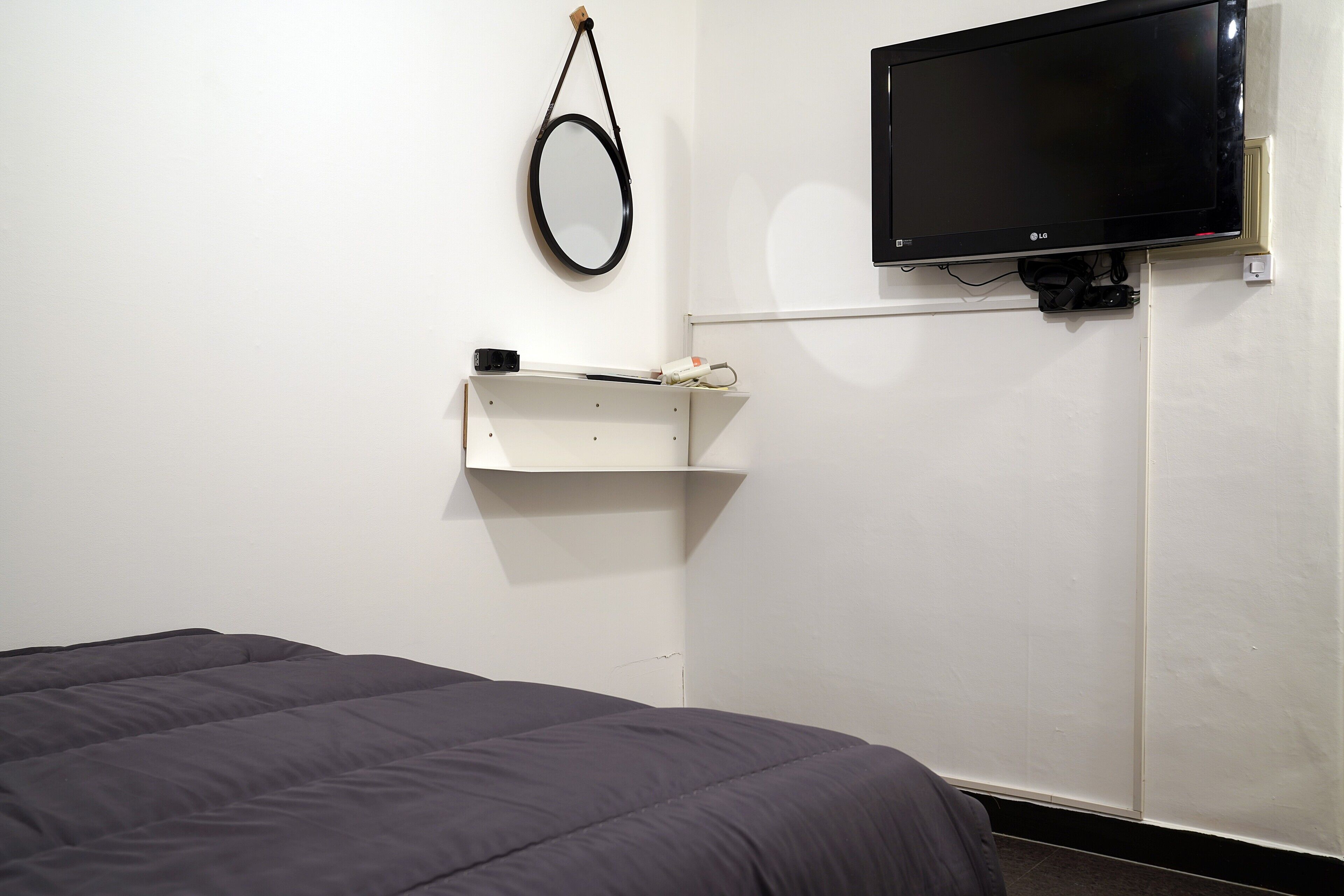 A room with a bed, a mounted TV, a small shelf, and a ring light.