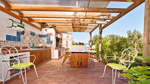 An outdoor patio with a wooden table and chairs, a tiled backsplash, and a view of greenery and mountains in the background. - Appartamento le Campanelle (San Felice del Benaco)