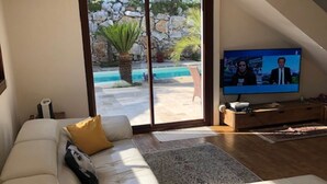 A modern living room with a white sofa, a flat-screen TV, and a view of an outdoor pool area.