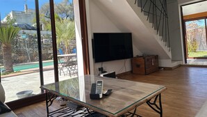 A spacious living area with a large glass table, a flat-screen TV mounted on the wall, and a view of a swimming pool outside.