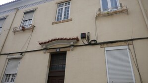 Fachada de edificio con entrada central, varias ventanas con contraventanas y un cuadro eléctrico visible.