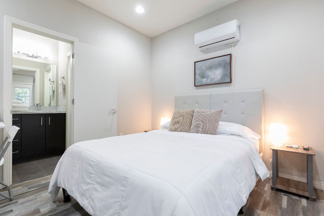 A bedroom with a large bed, a bedside table with a lamp, an air conditioning unit, and a framed picture on the wall.