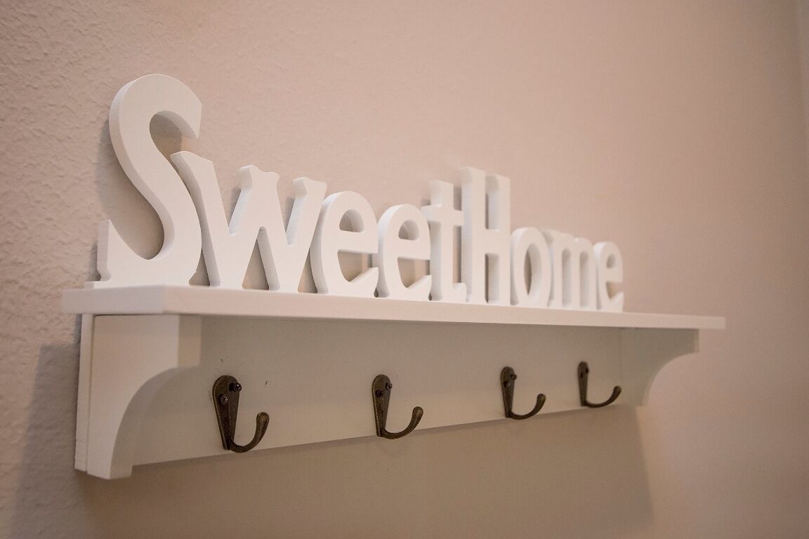 Uma prateleira branca com a frase "Sweet Home" em letras grandes. Abaixo da prateleira, há três ganchos.