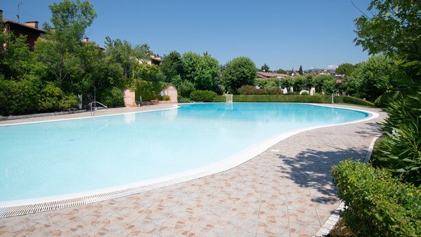 A swimming pool with a tiled deck, surrounded by greenery and houses in the background.
