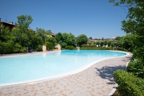 Ein Schwimmbecken mit Fliesenbord und umgebender Bepflanzung, im Hintergrund Wohnhäuser.