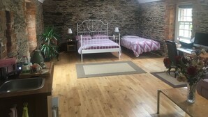 Une chambre avec un plafond en pente, des murs en pierre, un cadre de lit en métal blanc, un bureau avec un ordinateur et une fenêtre.