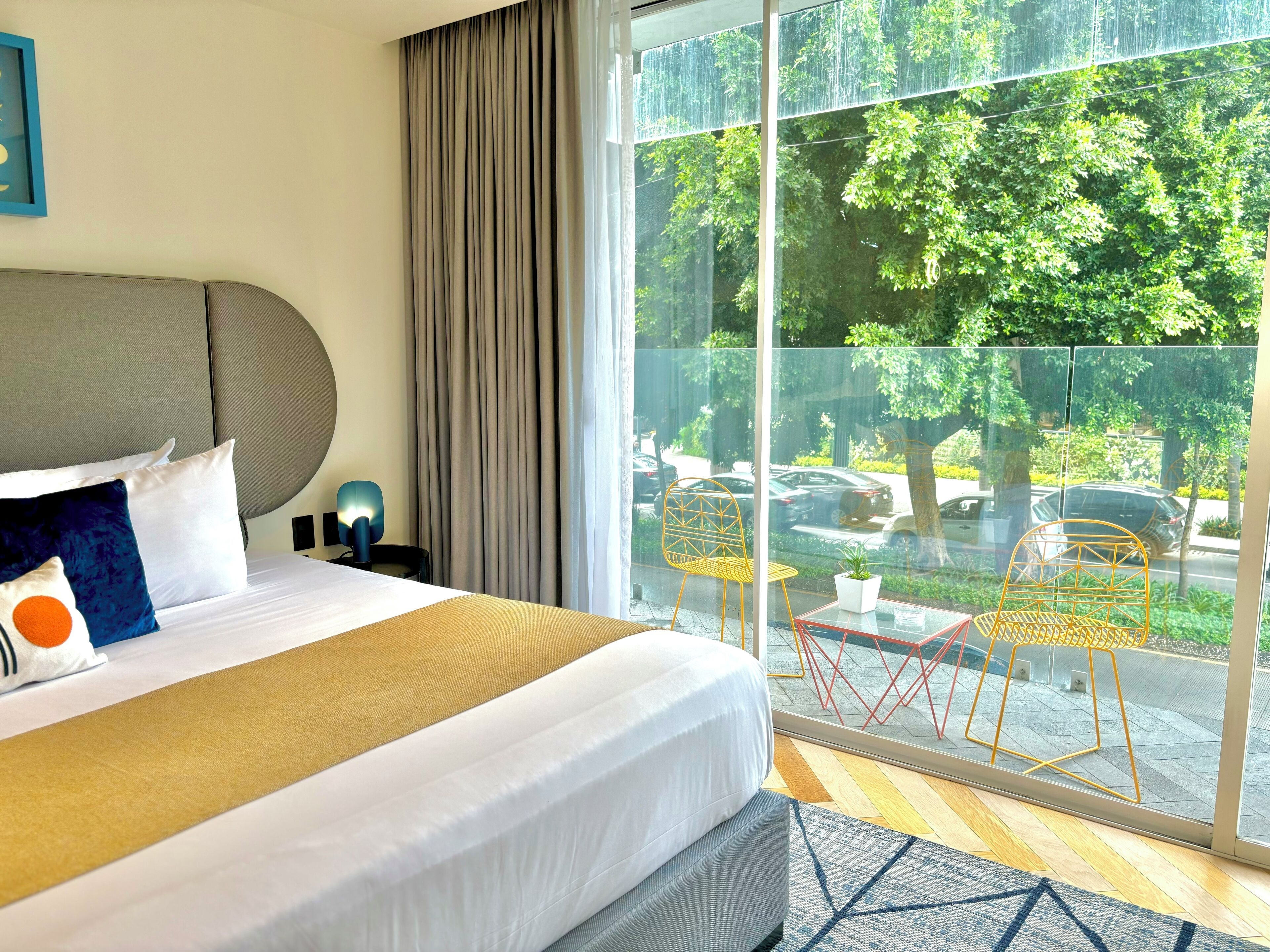 Ein Hotelzimmer mit einem großen Bett, Blick auf die Straße durch eine Glaswand und einem kleinen Tisch mit Stühlen draußen.