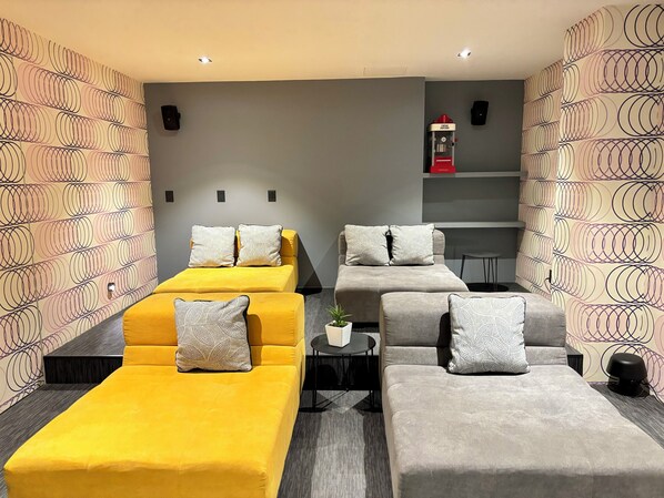 Habitación de hotel con dos camas, una amarilla y otra gris, ambas con almohadones estampados. Hay una mesita con una planta y un objeto rojo en un estante sobre las camas.