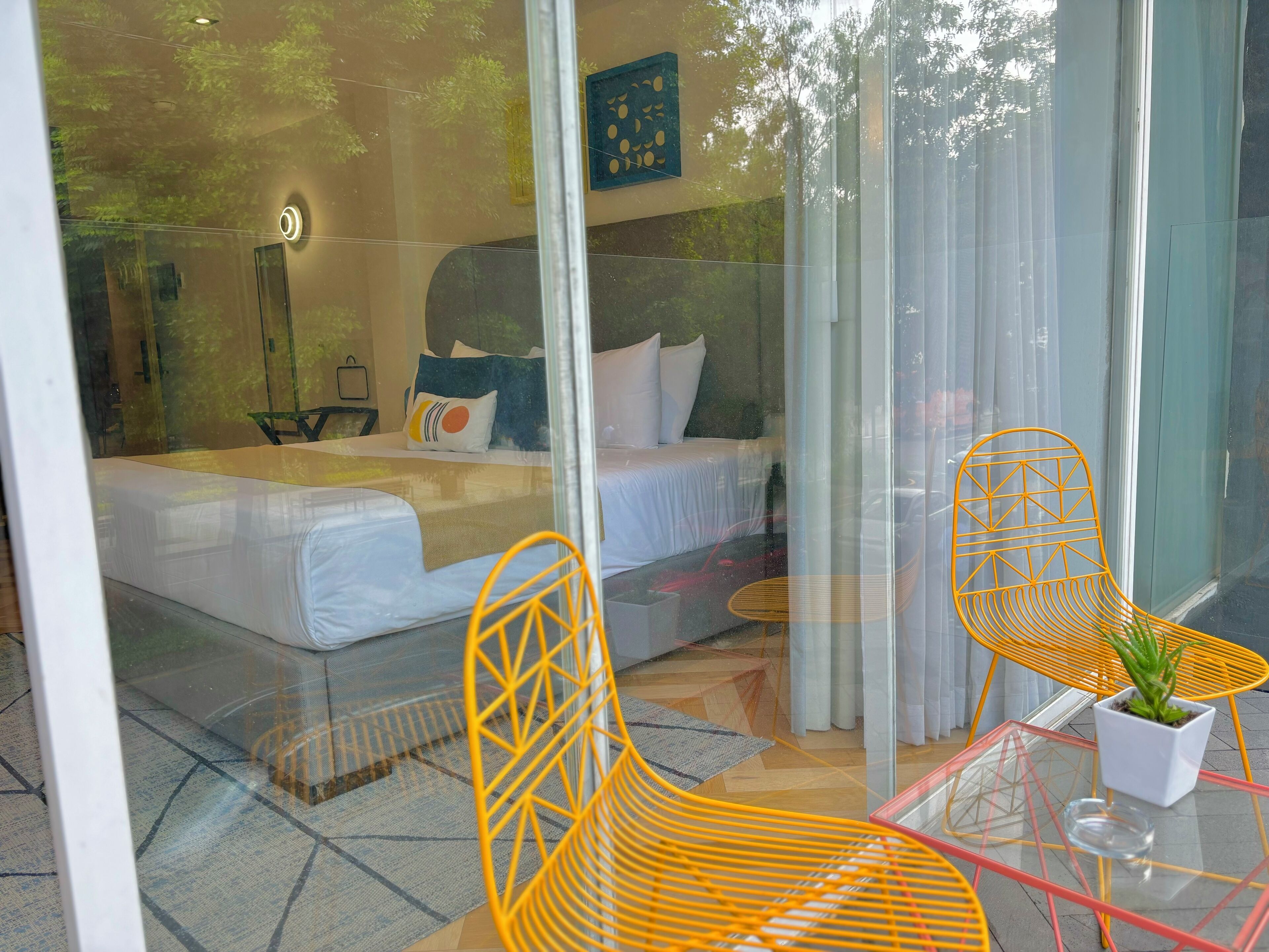 A modern bedroom with a glass partition, a bed with white linens, a yellow chair, and a small table with a plant.