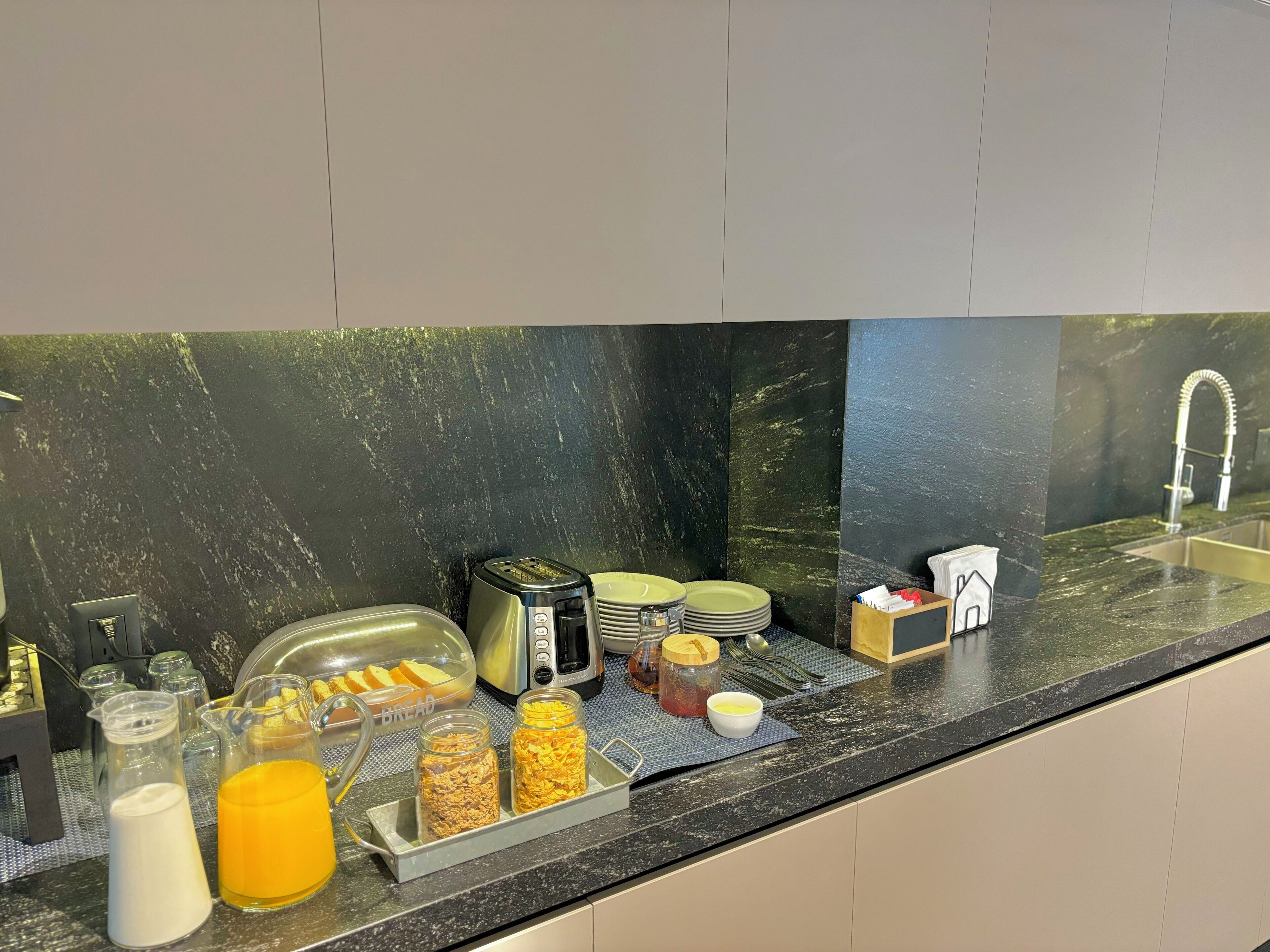 A breakfast buffet setup with a toaster, plates, a bread basket, and various food items on a marble countertop.