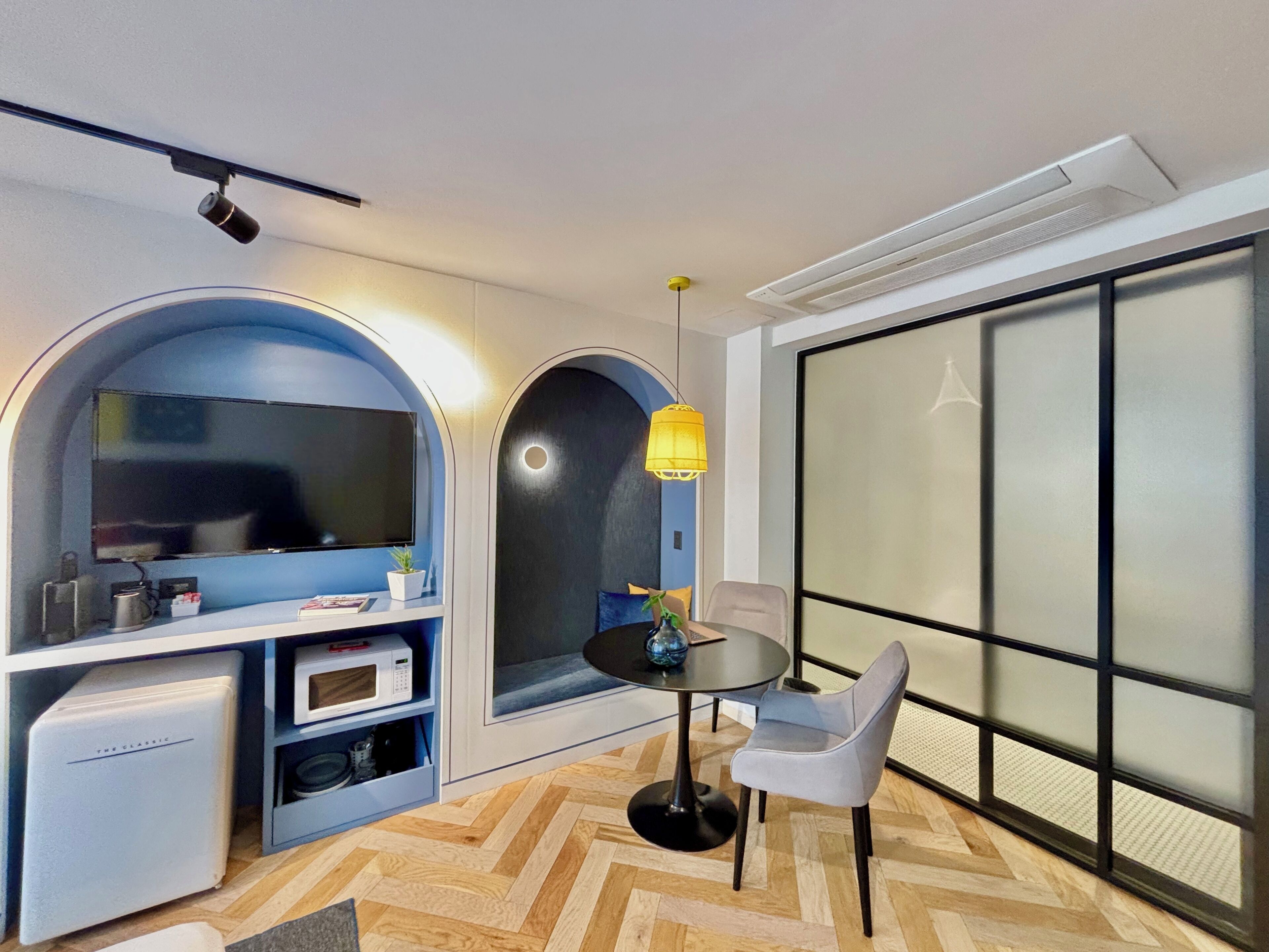 Una sala moderna con televisor, microondas y unidad de almacenamiento. Hay una mesa redonda con una silla, una lámpara de pie y un ventanal con vistas amplias.