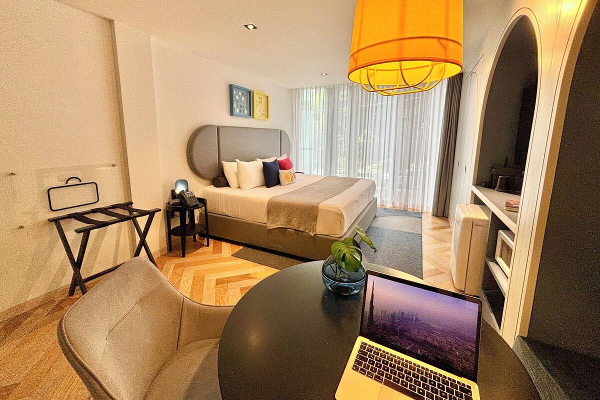 Habitación de hotel moderna con una cama, un escritorio con una computadora portátil, una silla y una pequeña mesa con una planta.