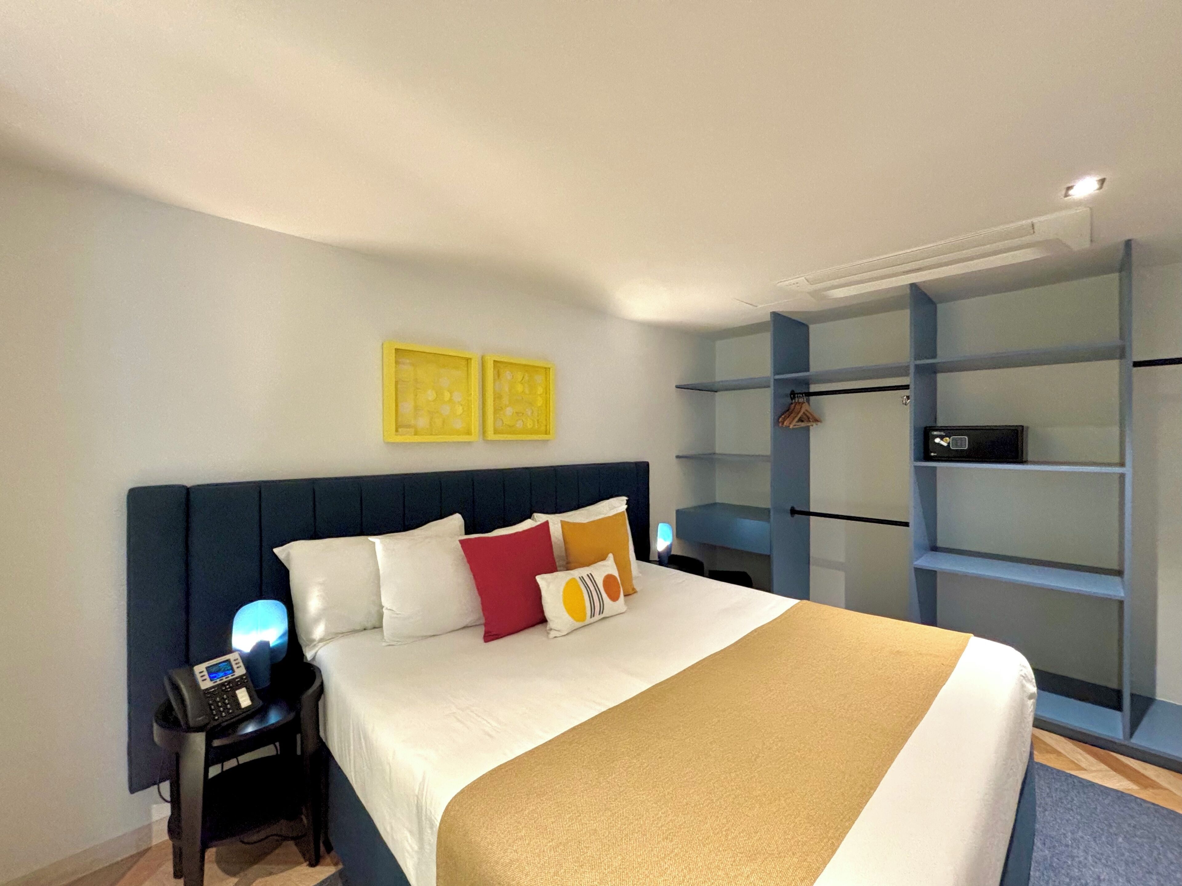 A modern hotel room with a large bed, a bedside table with a phone, a wardrobe with shelves, and two framed pictures on the wall.