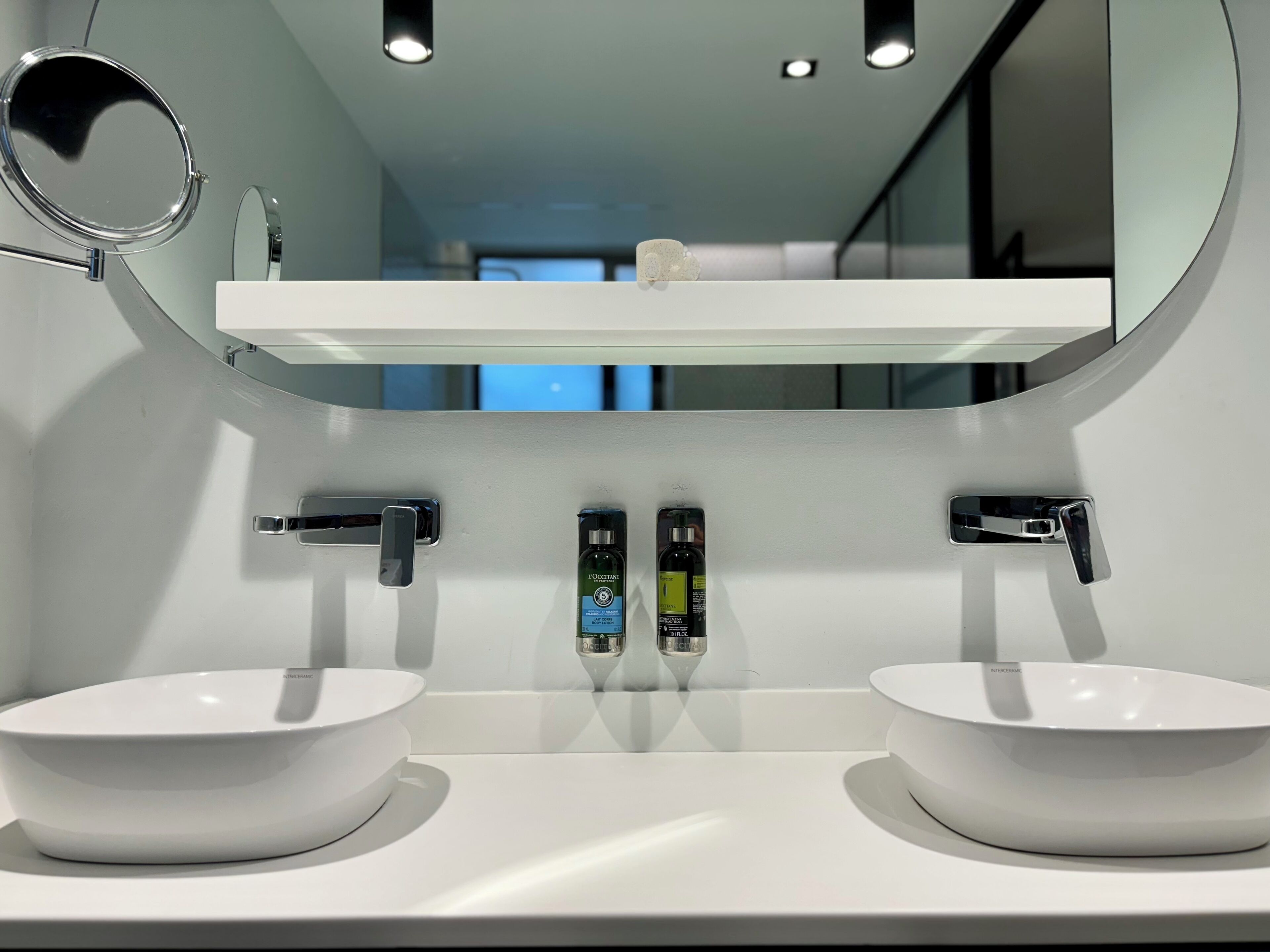 A modern bathroom with two white sinks, a mirror, and a shelf above the sinks.