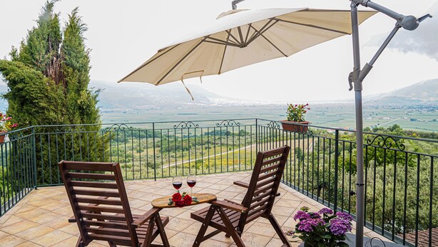 Een balkon met twee houten stoelen, een tafel en een grote parasol. Er staan planten in potten en er is uitzicht op het landschap.