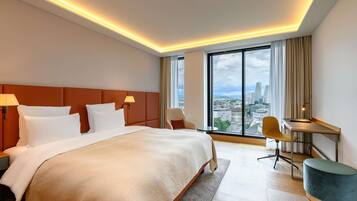 A modern hotel room with a large bed, a desk, a chair, and a view of the cityscape.