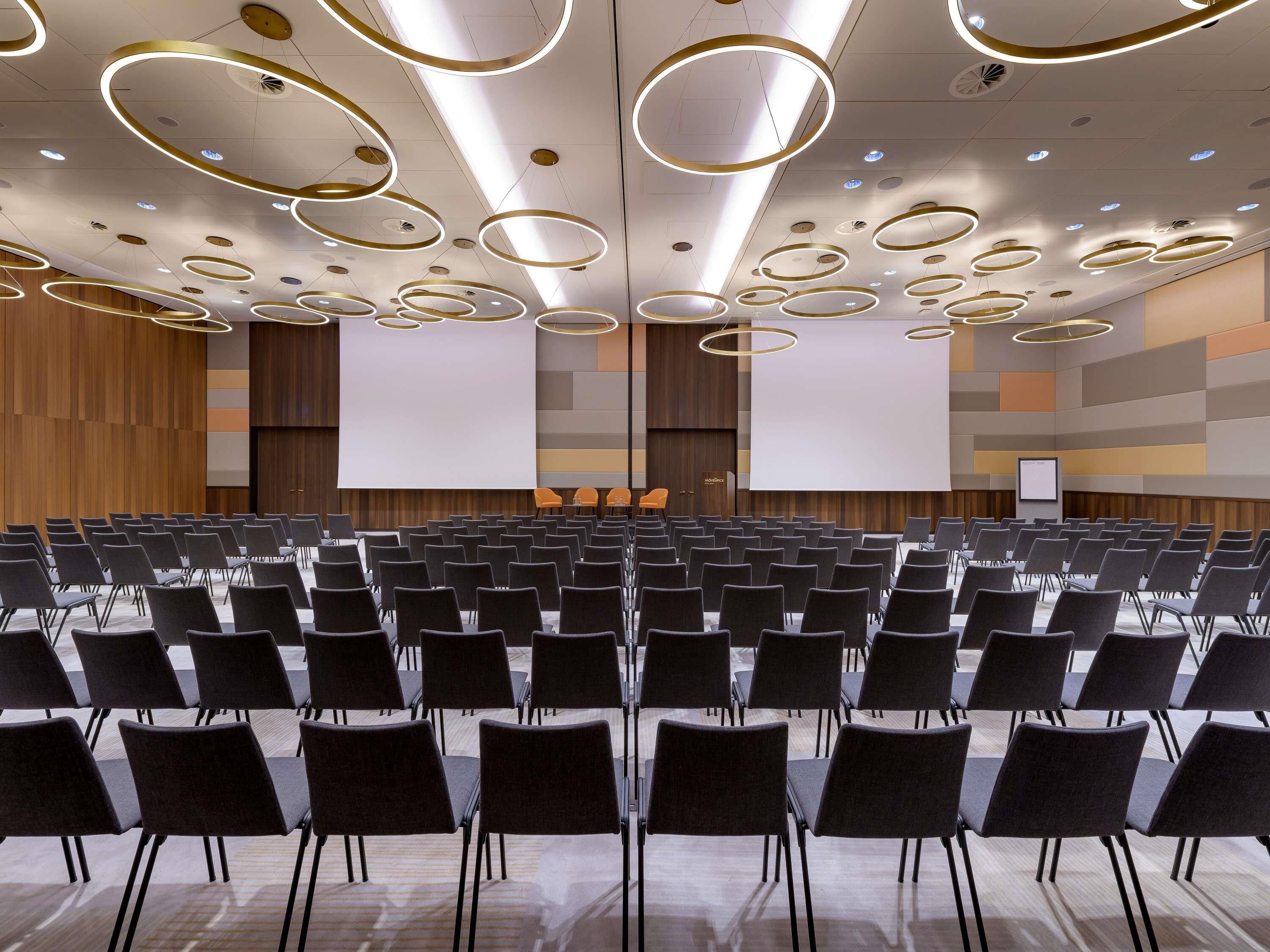 Un salón de conferencias con filas de sillas, una pantalla grande y modernos apliques circulares en el techo.