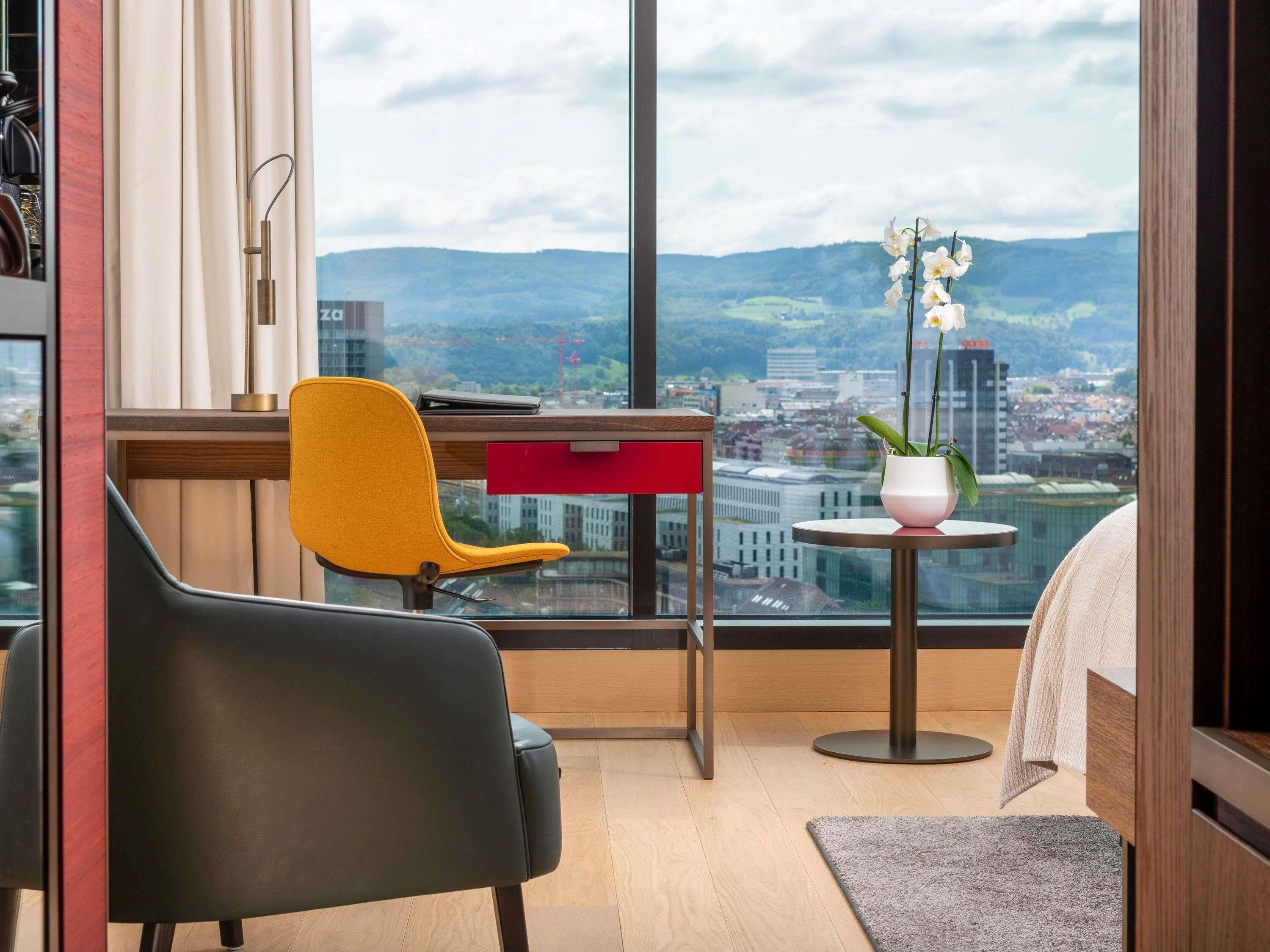 A modern room with a city view, featuring a desk, two chairs, a small table, and a potted orchid.