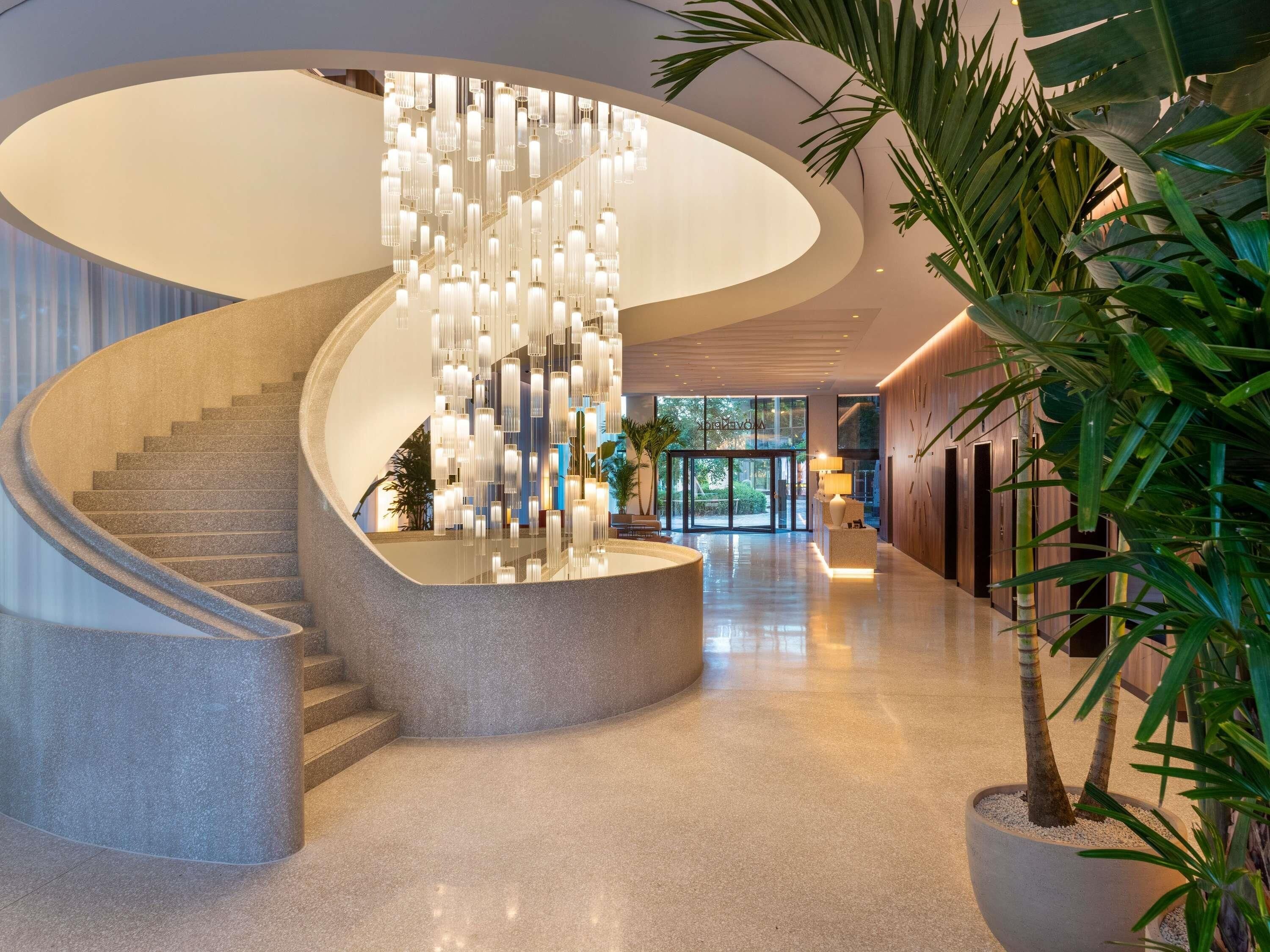 Un hall d’hôtel moderne avec un escalier en colimaçon, un lustre central et un comptoir d’accueil.