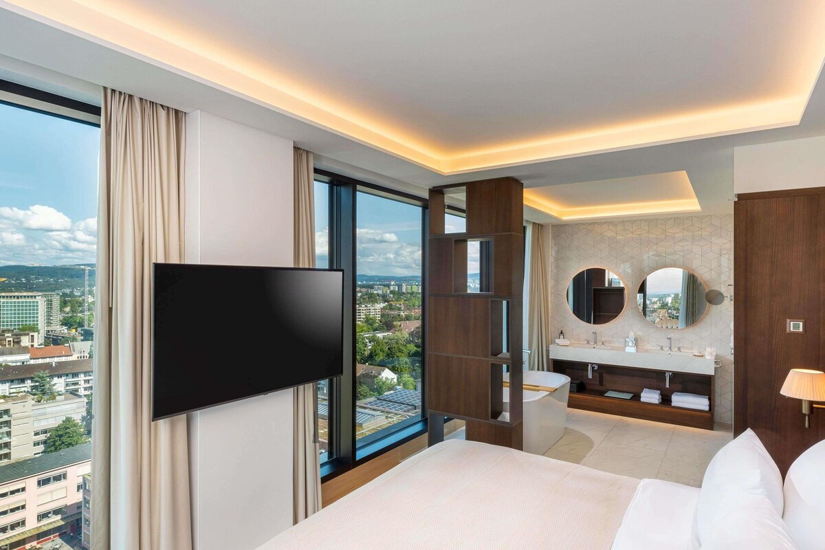 A hotel room with a large bed, a TV, a bathroom with a mirror, and a city view.