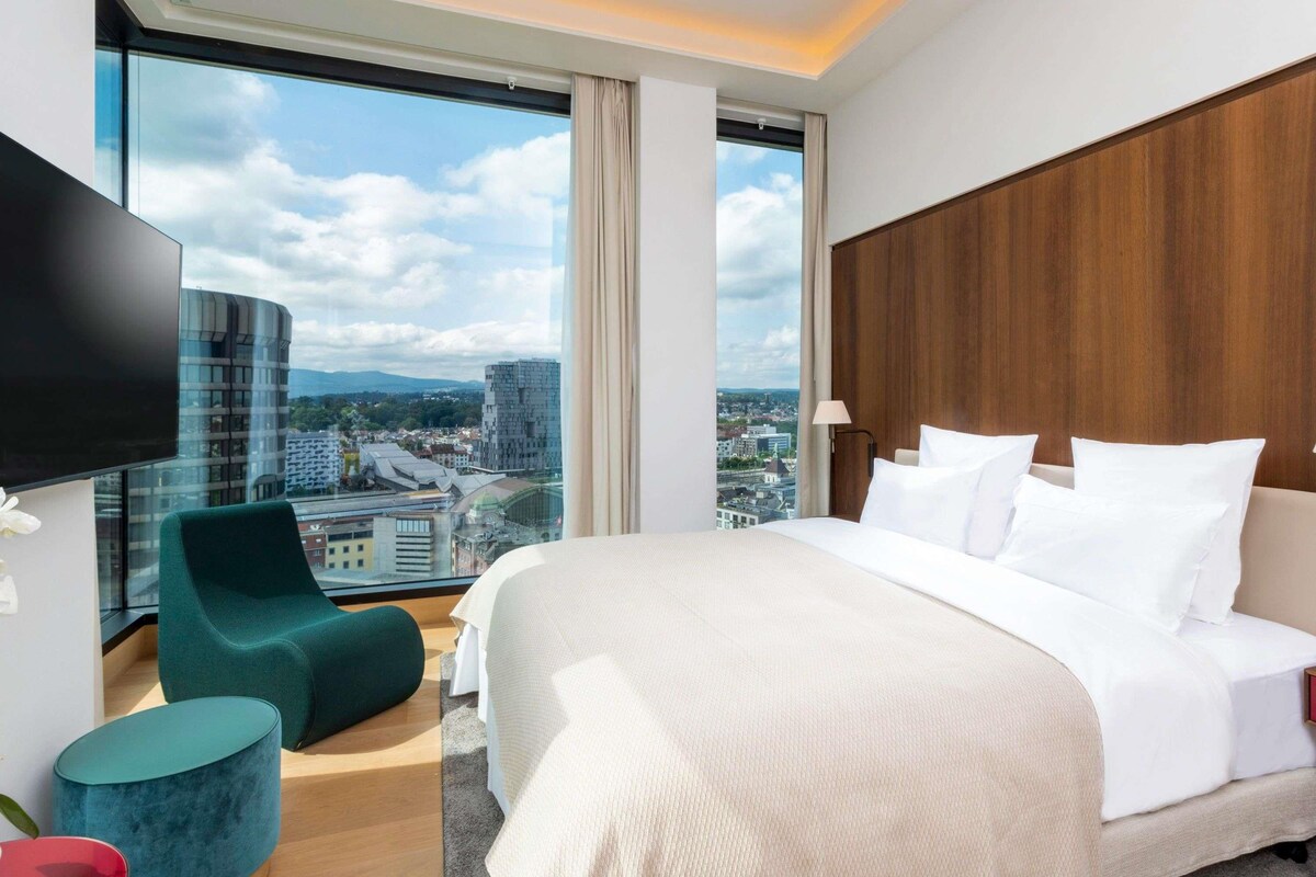 A modern hotel room with a large bed, a television, a city view, and a wooden headboard.
